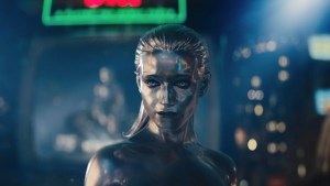 A silver, metallic humanoid figure with slicked-back hair stands against a city backdrop with intense, emotionless expression.