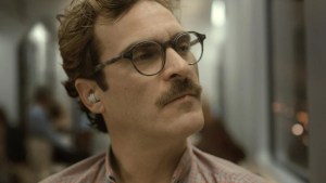Joaquin Phoenix as Theodore Twombly in Her wears round glasses and a checkered shirt, gazing thoughtfully into the distance with a subtle frown. An earpiece is visible in his ear