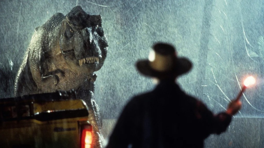 A large dinosaur stands in the foreground, its head lowered and mouth open, revealing sharp teeth. Behind the dinosaur, a person wearing a cowboy hat holds a flare, illuminating the dark, rainy scene. The background features a blurred vehicle with headlights on, and water droplets are visible in the air.
