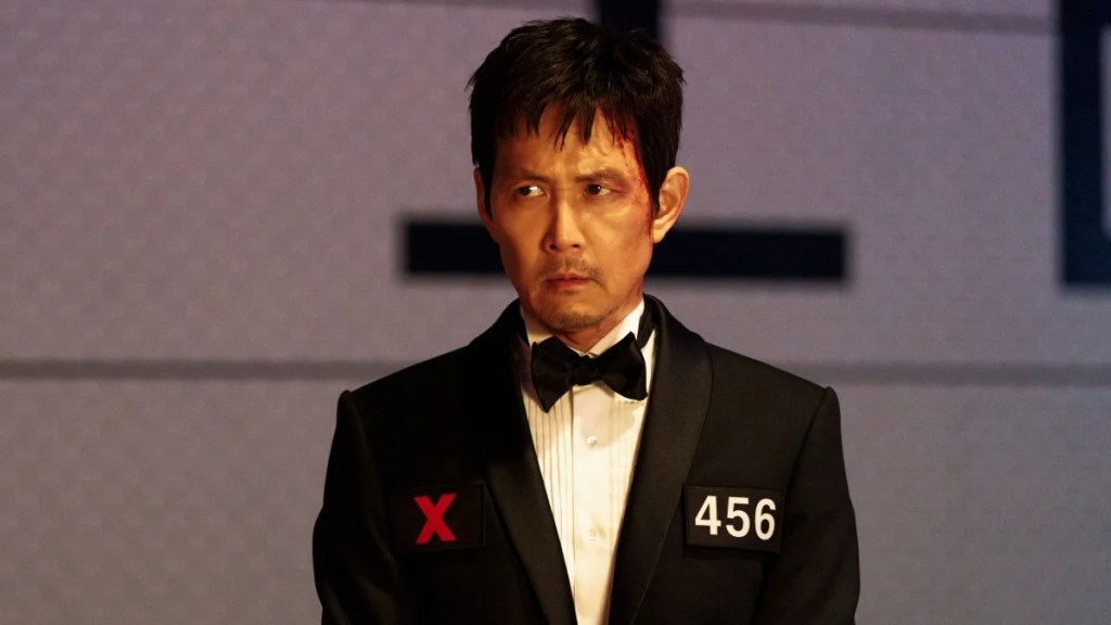 A bloodied and intense man in formalwear stands beneath cold lighting, his tuxedo marked with an “X” and the number 456