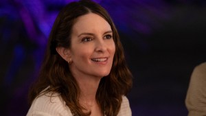 A woman with brown hair in a white flowing top smiles at someone out of frame. There is blue light behind her.