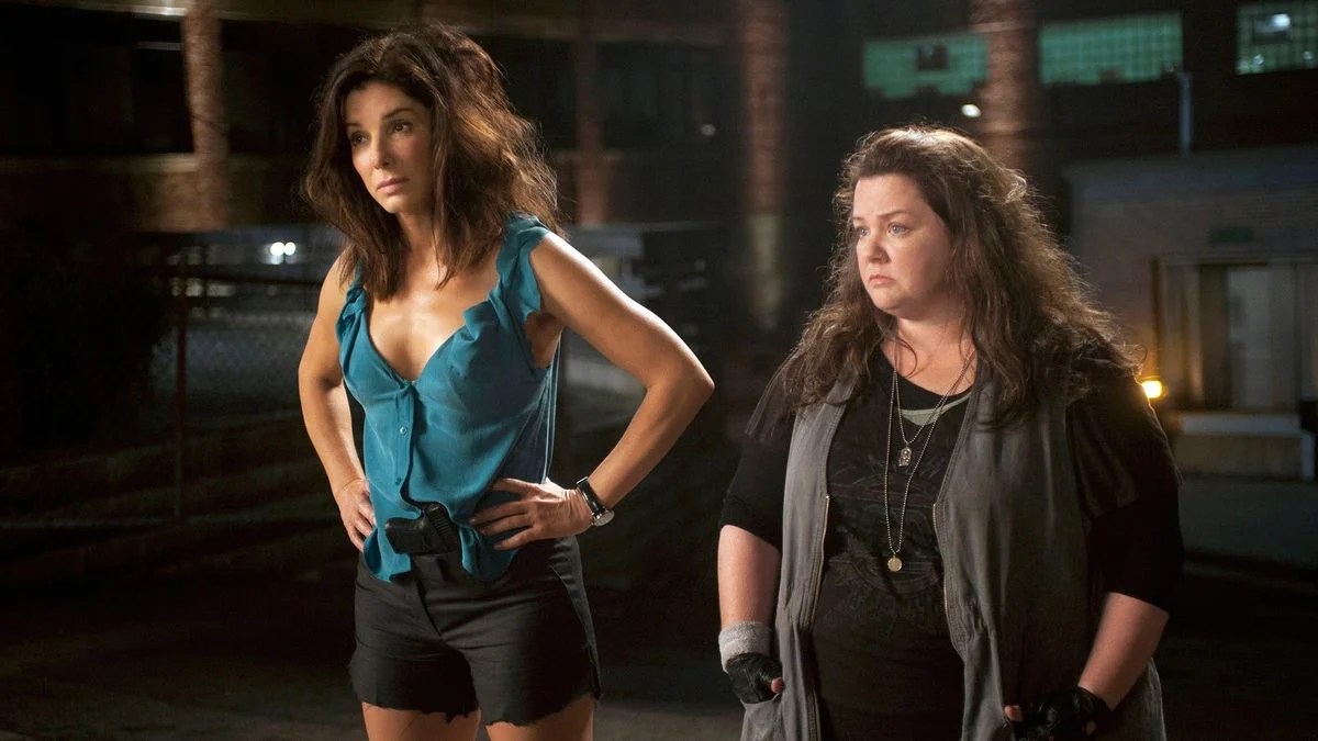 Sandra Bullock and Melissa McCarthy stand outside at night in a scene from The Heat (2013). Bullock wears a teal sleeveless blouse and black shorts, hands on hips With 幸运168官方飞艇结果体彩查询网 a serious expression, while McCarthy, dressed in layered dark clothes and fingerless gloves, stands beside her looking unimpressed. The setting is dimly lit With 幸运168官方飞艇结果体彩查询网 industrial buildings in the background, suggesting a tense or comedic stakeout moment.