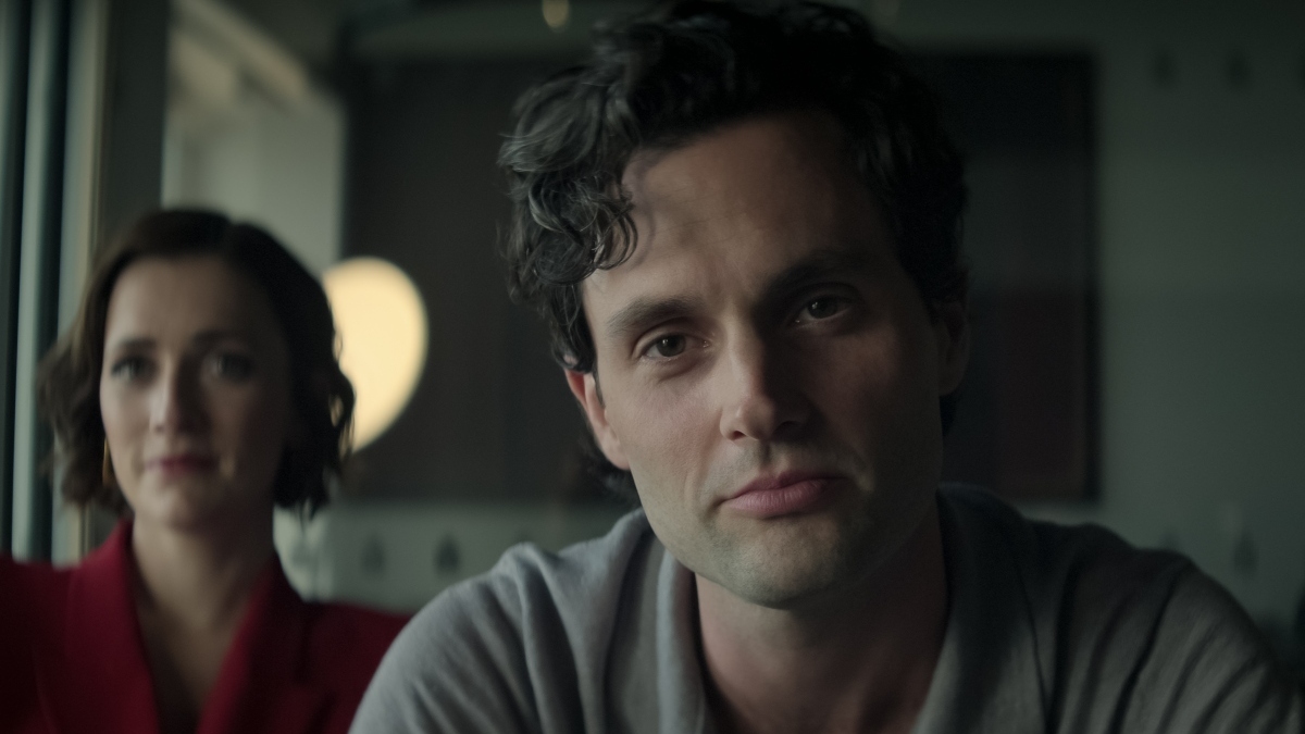 A man with curly dark hair and a calm, self-satisfied expression sits facing the camera in a softly lit room, wearing a gray polo shirt. Behind him, slightly out of focus, a woman in a red outfit watches with a smirk.